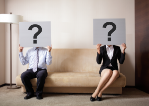 Man and woman sitting on opposite sides of the couch holding up a question mark card in front of their face.